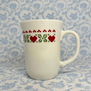 May include: White ceramic mug with a red and green embroidered design of hearts and flowers around the top.