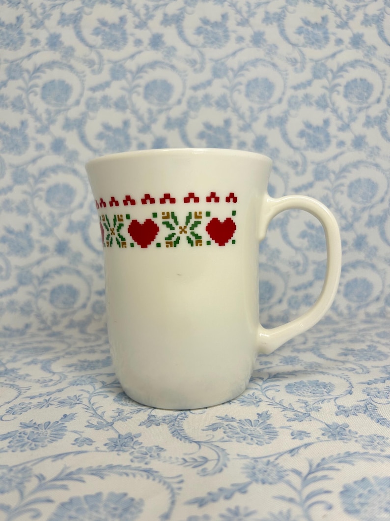 May include: White ceramic mug with a red and green embroidered design of hearts and flowers around the top.