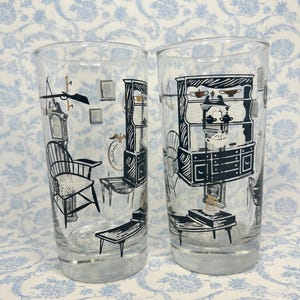 May include: Two clear drinking glasses with a silver base, each featuring a black and white illustration of a room interior. The illustrations include furniture, clocks, and decorative elements, creating a vintage aesthetic.