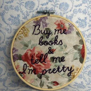 May include: A circular wooden embroidery hoop framing fabric with a floral pattern in shades of red, yellow, and green. The text "Buy me books & tell me I'm pretty" is embroidered in dark purple thread across the center of the floral fabric.