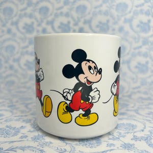 May include: White ceramic mug with a repeating design of Mickey Mouse in a running pose. Mickey is shown with black ears, a black body, red shorts, and yellow shoes. The mug has a cylindrical shape and a smooth surface.