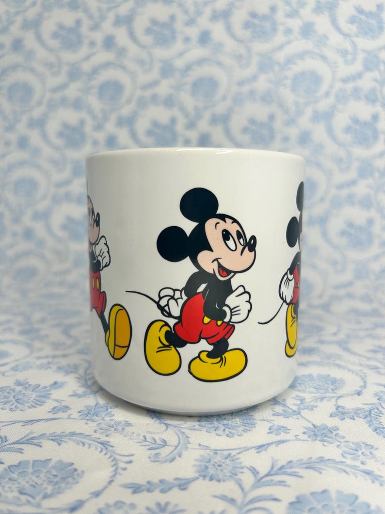 May include: White ceramic mug with a repeating design of Mickey Mouse in a running pose. Mickey is shown with black ears, a black body, red shorts, and yellow shoes. The mug has a cylindrical shape and a smooth surface.
