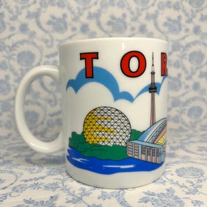 May include: White ceramic mug with the word "TORONTO" in red letters. The mug features illustrations of Toronto landmarks, including the CN Tower, a geodesic dome, and a stadium. The mug has a white handle and a blue and white floral background.