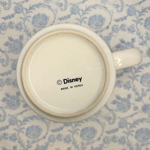 May include: White ceramic mug with a handle, featuring the Disney logo and the text "MADE IN KOREA" on the base. The mug is set against a light blue floral patterned background.
