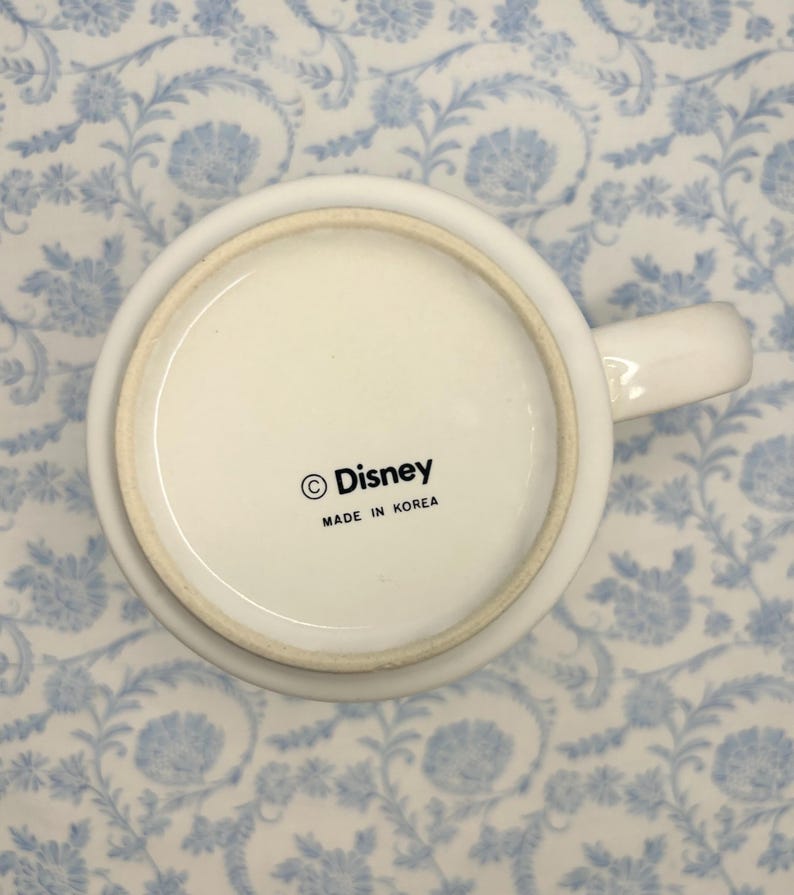 May include: White ceramic mug with a handle, featuring the Disney logo and the text "MADE IN KOREA" on the base. The mug is set against a light blue floral patterned background.
