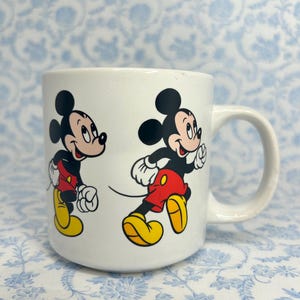 May include: White ceramic mug with two images of Mickey Mouse running. Mickey is shown in his iconic outfit: red shorts, yellow shoes, white gloves, and black ears. The mug has a curved handle.