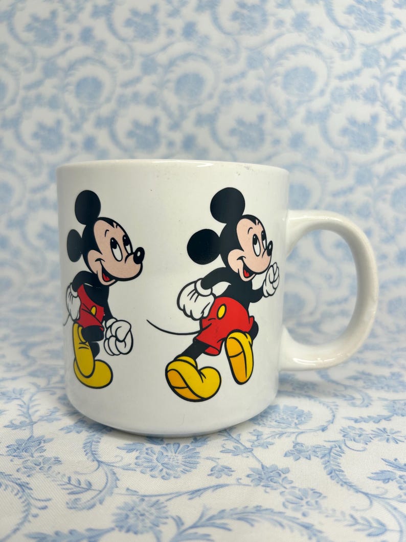 May include: White ceramic mug with two images of Mickey Mouse running. Mickey is shown in his iconic outfit: red shorts, yellow shoes, white gloves, and black ears. The mug has a curved handle.