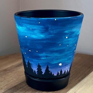May include: A hand-painted ceramic flower pot with a night sky design. The pot features a dark blue base and rim, with a gradient of blues and white stars painted on the exterior. Silhouetted trees are at the bottom.