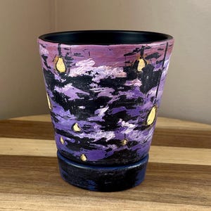 May include: Hand-painted ceramic pot with a night sky design. The pot features a black rim and base, with a body painted in shades of purple, black, and white, with yellow accents. The design resembles a starry night scene, perfect for holding plants.