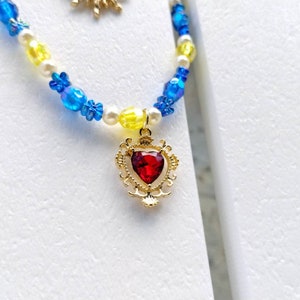 Peut inclure: Un pendentif en forme de cœur doré avec une pierre précieuse rouge pend d'un collier de perles. Le collier est composé de perles bleues, jaunes et blanches.