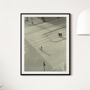 May include: Black framed art print featuring an aerial view of a city street scene. The black and white photograph shows a person on a bicycle and two people standing on a corner. The print is displayed on a white wall.