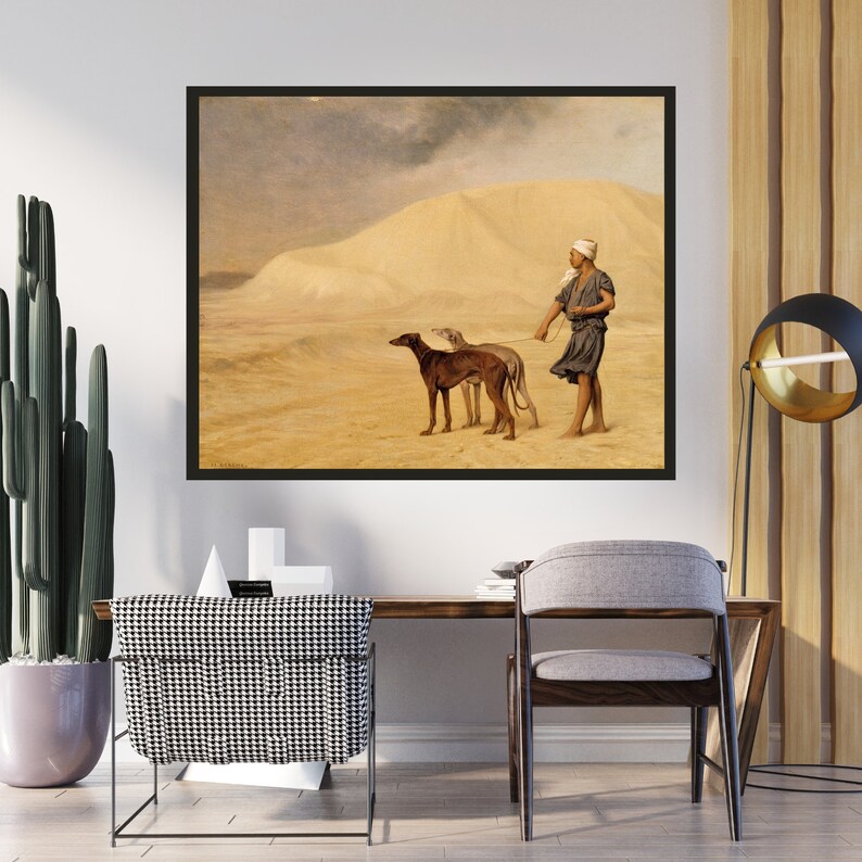 Op de afbeelding: Ingelijste prent van een woestijnsc&egrave;ne. Een persoon leidt twee honden aan de lijn door een zanderig landschap. Het kunstwerk toont een figuur met een hoofdbedekking en donkere kleding. De honden zijn bruin en grijs. Het kunstwerk zit in een zwarte lijst.