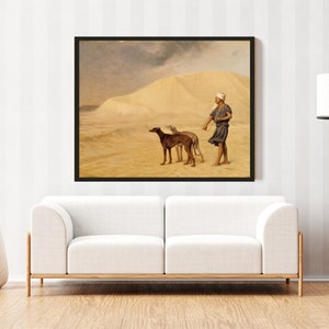Op de afbeelding: Een ingelijste prent van een antiek schilderij dat een woestijnlandschap toont. Een persoon in traditionele kleding loopt met twee honden aan de lijn. De achtergrond toont een zandduin en een bewolkte lucht. Het kunstwerk wordt boven een witte bank getoond.