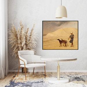 Op de afbeelding: Ingelijste kunstdruk met een woestijnlandschap met een persoon en twee honden. Het kunstwerk toont een persoon in traditionele kleding die een riem vasthoudt, met twee slanke honden. De achtergrond toont een zandduin onder een bewolkte hemel. De lijst is zwart.