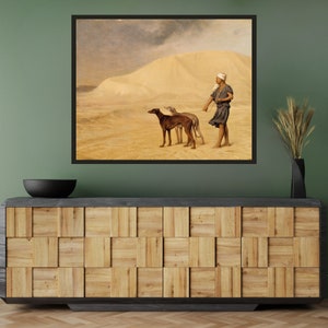 Op de afbeelding: Ingelijste kunstdruk met een woestijnlandschap met een persoon en twee windhonden. Het kunstwerk toont een zanderige omgeving, een bewolkte lucht en een figuur in traditionele kleding. De print wordt boven een houten kast getoond.