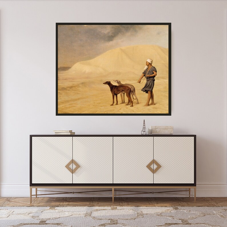 Op de afbeelding: Een ingelijste prent van een woestijnsc&egrave;ne met een persoon en twee honden. Het kunstwerk toont een zanderig landschap met een grote zandduin op de achtergrond. De honden zijn bruin en grijs, en de persoon draagt een hoofdbedekking en een donkere tuniek.