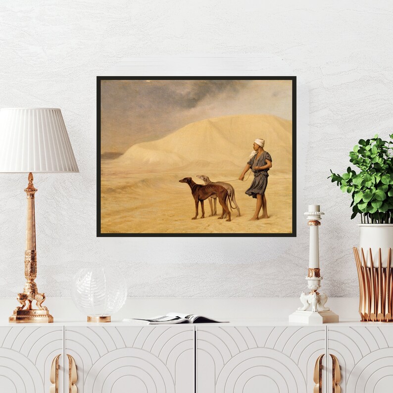 Op de afbeelding: Ingelijste print van een woestijnlandschap met een persoon en twee honden. De persoon, gekleed in een hoofdbedekking en tuniek, leidt de honden over het zand. De honden zijn bruin en grijs. De lucht is bewolkt.