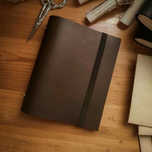 May include: A dark brown leather journal with a black elastic band closure. The journal is closed and lying on a wooden surface. A pair of silver scissors and rolled-up scrolls are also visible.