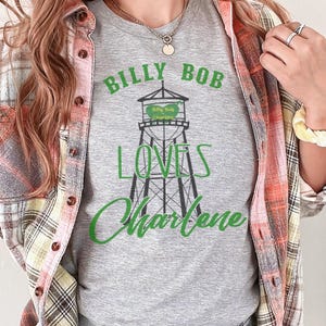 May include: A gray t-shirt with the text "BILLY BOB LOVES Charlene" in green. The design features a water tower graphic with the words "Billy Bob Charlene" on the water tank. The person is wearing a plaid shirt.
