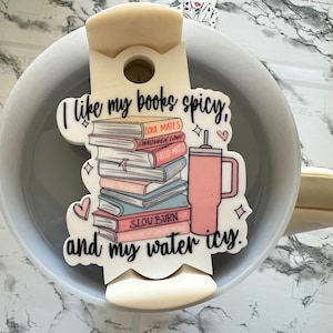 May include: A white ceramic mug with a white and pink sticker that says "I like my books spicy, and my water icy." The sticker features a stack of books with titles like "Soul Mates", "Forbidden Love", "Fated Mates", and "Slow Burn". A pink tumbler with a straw is next to the books.