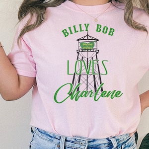 May include: Pink t-shirt with a graphic of a water tower that says "Billy Bob Loves Charlene" in green text.
