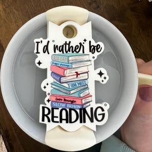 Peut inclure: Une tasse en céramique blanche avec une anse crème et une étiquette décorative avec une pile de livres. L'étiquette indique "I'd rather be READING". La tasse est tenue par une personne.