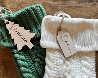 Op de afbeelding: Twee gebreide kerstsokken met gepersonaliseerde houten labels. De groene sok heeft een label in de vorm van een boom met de naam "Duncan" en de witte sok heeft een rechthoekig label met de naam "Emma".