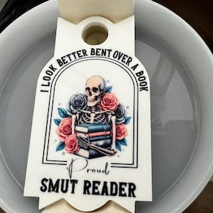 May include: A white ceramic mug with a decorative tag. The tag features a skeleton with roses and books, and the text "I LOOK BETTER BENT OVER A BOOK" and "PROUD SMUT READER".