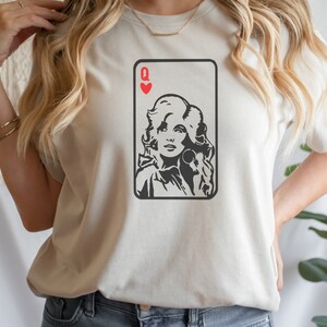 Peut inclure: T-shirt cr&egrave;me avec un graphisme noir et blanc d'un portrait de femme dans un motif de carte &agrave; jouer. La carte est la Dame de C&oelig;ur, avec un symbole de c&oelig;ur rouge en haut. Le t-shirt a des manches courtes.