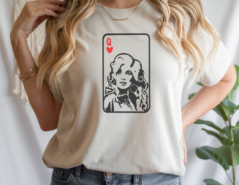 Peut inclure: T-shirt cr&egrave;me avec un graphisme noir et blanc d'un portrait de femme dans un motif de carte &agrave; jouer. La carte est la Dame de C&oelig;ur, avec un symbole de c&oelig;ur rouge en haut. Le t-shirt a des manches courtes.