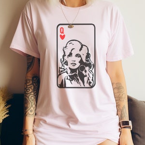 Peut inclure: T-shirt rose clair avec un motif de carte &agrave; jouer en noir et blanc. La carte repr&eacute;sente la Reine de C&oelig;ur, avec un portrait de femme aux cheveux boucl&eacute;s et boucles d'oreilles. Le t-shirt a un col rond et des manches courtes.