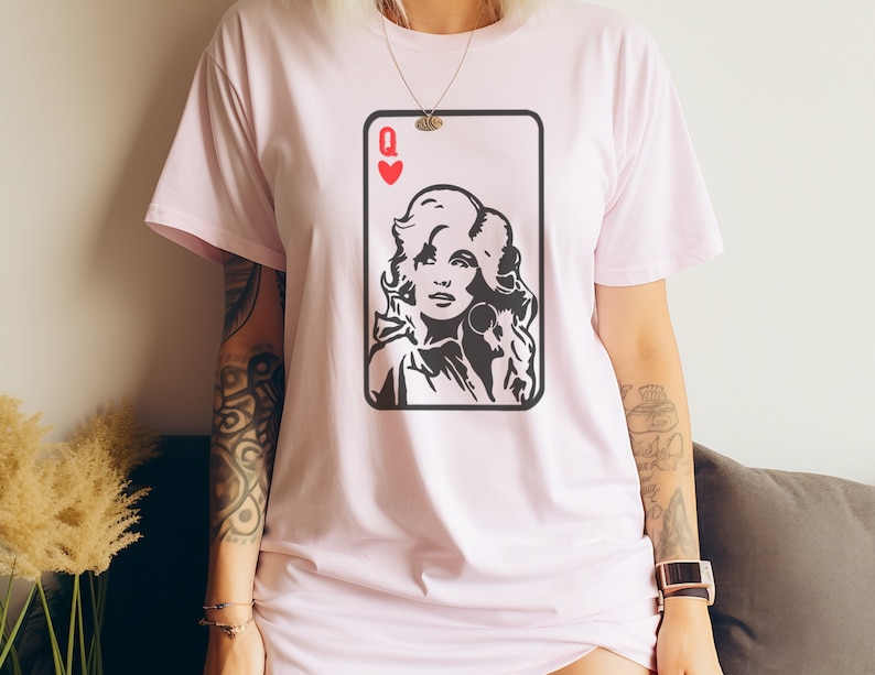 Peut inclure: T-shirt rose clair avec un motif de carte &agrave; jouer en noir et blanc. La carte repr&eacute;sente la Reine de C&oelig;ur, avec un portrait de femme aux cheveux boucl&eacute;s et boucles d'oreilles. Le t-shirt a un col rond et des manches courtes.