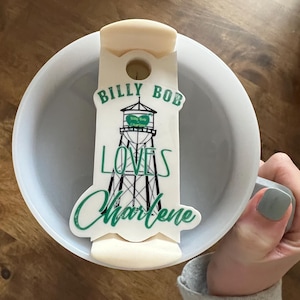Może przedstawiać: Biały kubek ceramiczny z białą silikonową pokrywką. Pokrywka ma zielono-czarną grafikę wieży ciśnień z tekstem "Billy Bob Loves Charlene".
