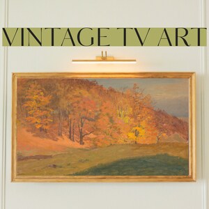 May include: A framed painting of a landscape with a hillside covered in autumn trees. The trees are a mix of orange, yellow, and red. The painting is in a gold frame and is hanging on a white wall.
