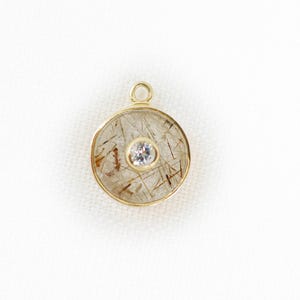 May include: A gold pendant with a round, clear stone center. The stone has a smaller, clear stone in the middle. The pendant has a gold loop at the top for hanging. The pendant is set against a white background.