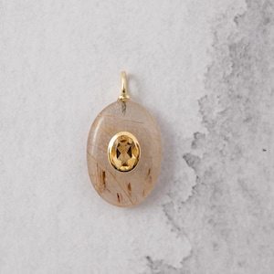 May include: An oval-shaped pendant featuring a translucent, light brown stone with dark inclusions. A small, oval-cut yellow gemstone is bezel-set in gold in the center of the larger stone. A gold bail is attached to the top of the pendant.