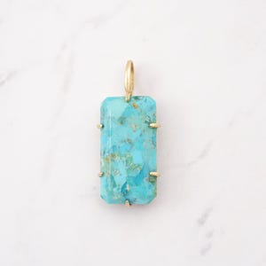 May include: A rectangular turquoise pendant featuring natural blue and green veining, set in a gold-toned metal prong setting with a small gold-toned bail at the top for attachment to a chain or necklace, showcasing a polished finish.