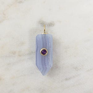 May include: A light blue gemstone pendant with a gold bail and a small, round, purple gemstone set in a gold bezel. The pendant is set against a white marble background.