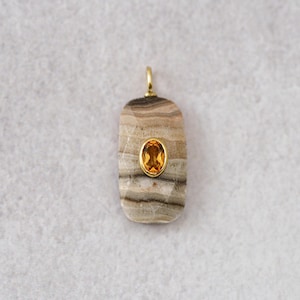 May include: A rectangular pendant with a gold bail and a gold-colored oval gemstone set in the center. The pendant is made of a light brown and gray striped stone.