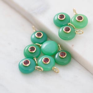 May include: A collection of green gemstone pendants with gold-colored loops and small red center stones. The pendants are round and have a polished finish, resting on a white surface.