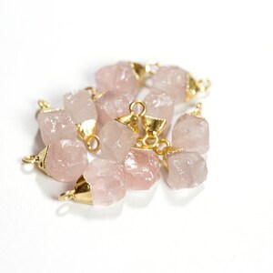 May include: A group of pink rose quartz crystal charms with gold-toned jump rings. The charms are irregularly shaped and have a natural, raw look.
