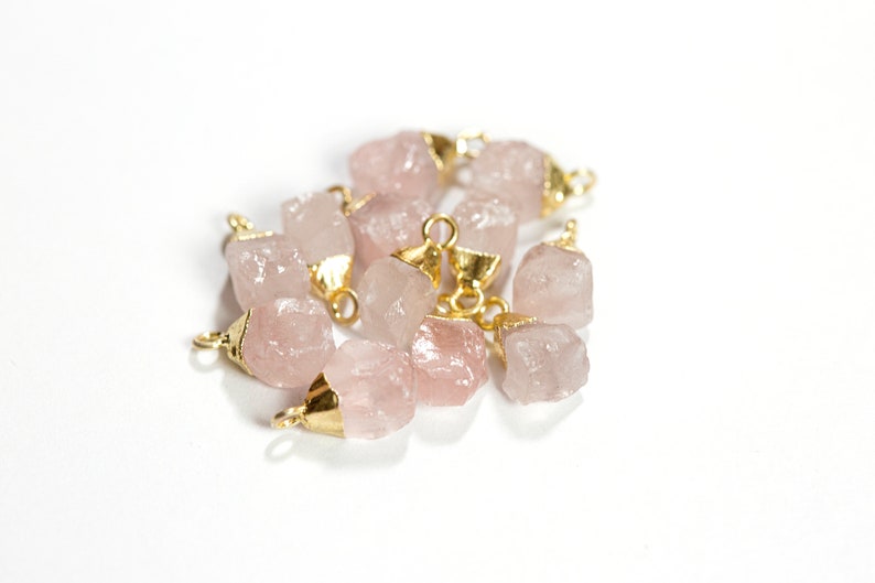 May include: A group of pink rose quartz crystal charms with gold-toned jump rings. The charms are irregularly shaped and have a natural, raw look.