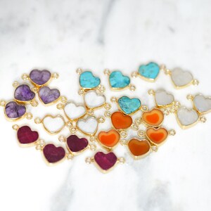 Pu&ograve; includere: Assortimento di ciondoli a forma di cuore in pietra preziosa con accenti dorati. I ciondoli sono di vari colori, tra cui viola, turchese, arancione e bianco.