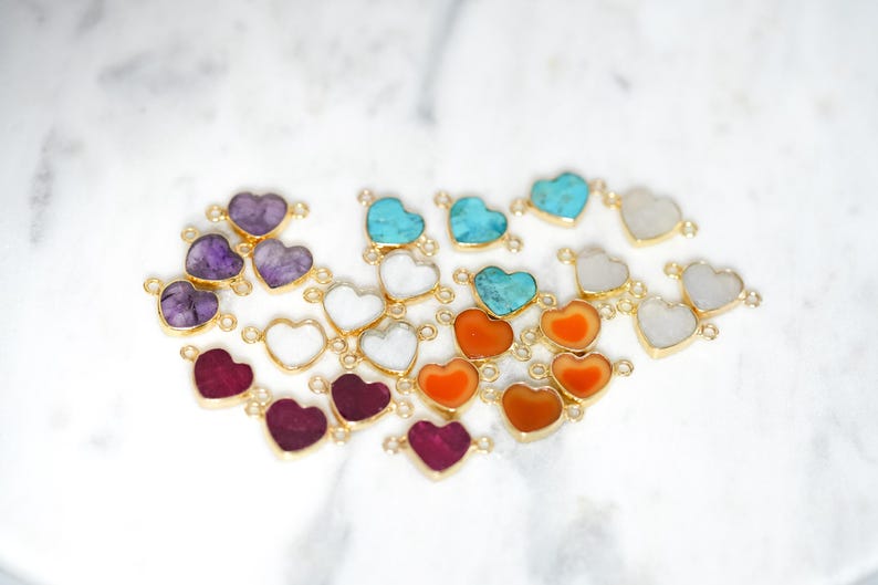 Pu&ograve; includere: Assortimento di ciondoli a forma di cuore in pietra preziosa con accenti dorati. I ciondoli sono di vari colori, tra cui viola, turchese, arancione e bianco.