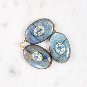 May include: Three oval-shaped pendants with a blue and gray iridescent stone and a smaller, oval-shaped, light blue stone set in a gold-colored metal. The pendants are displayed on a white marble surface.
