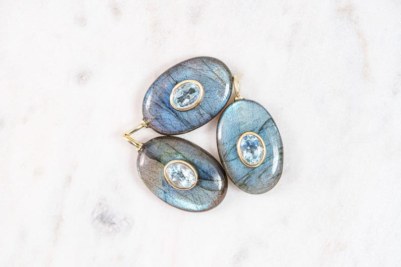 May include: Three oval-shaped pendants with a blue and gray iridescent stone and a smaller, oval-shaped, light blue stone set in a gold-colored metal. The pendants are displayed on a white marble surface.
