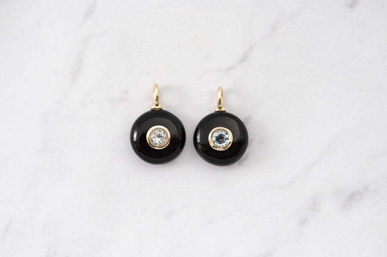 May include: Two circular pendants featuring a smooth black onyx base with a central bezel-set clear gemstone. Each pendant includes a small gold-toned metal bail at the top for attachment, creating a balanced and symmetrical jewelry design.