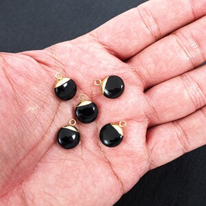 May include: Five black, round charms with gold-coloured accents and small loops. The charms are displayed on a hand, with a dark background. Suitable for jewellery making.