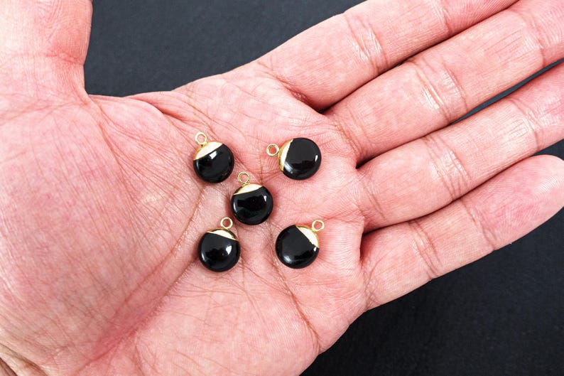 May include: Five black, round charms with gold-coloured accents and small loops. The charms are displayed on a hand, with a dark background. Suitable for jewellery making.