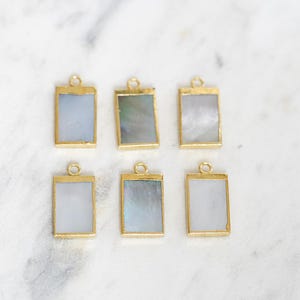 May include: Six rectangular pendants with gold-colored frames and iridescent centers. The pendants are arranged on a white and gray marble surface. Each pendant has a small loop at the top for attaching to a chain or other jewelry component.